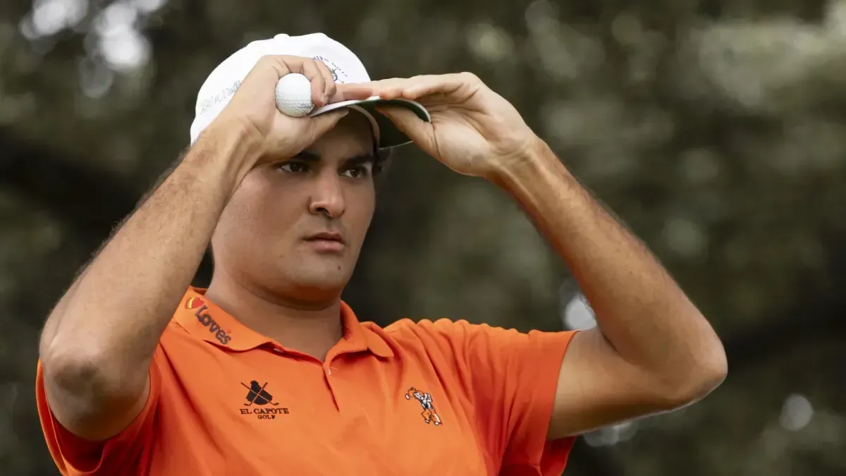 Golfista con polo naranja y gorra blanca sujeta una pelota junto a la frente y mira al horizonte entre árboles