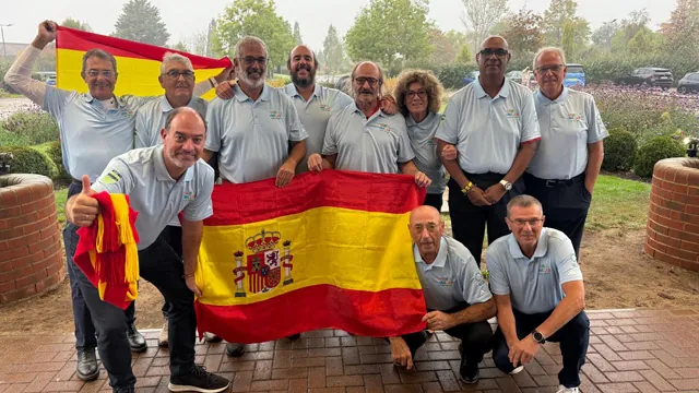 Grupo de personas con polos grises posando con banderas de España frente a un jardín con setos y macetas de ladrillo