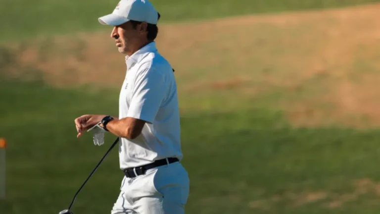 Hombre con gorra y polo blancos, con guante y palo de golf, caminando por un campo de hierba verde