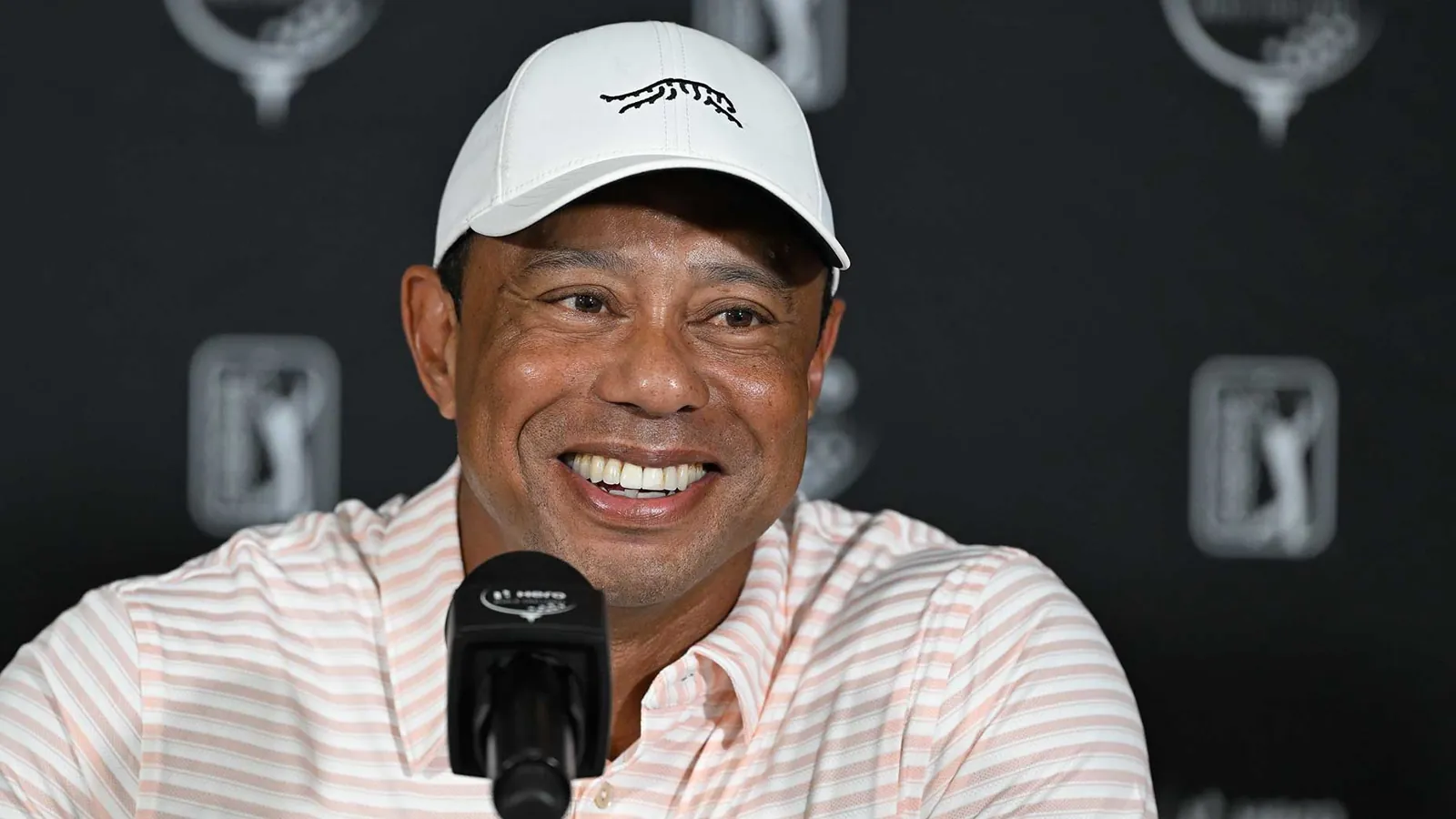 Golfista sonriente con gorra blanca y polo a rayas rosas, sentado en una rueda de prensa con micrófono y fondo oscuro.