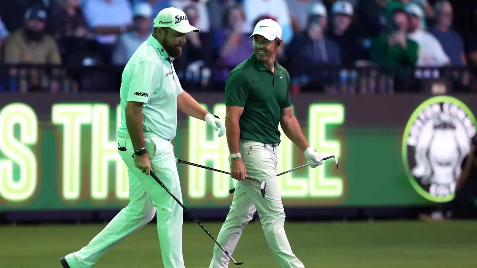 Dos golfistas, uno de blanco y otro de verde, caminan sonriendo con los palos en la mano ante un cartel luminoso