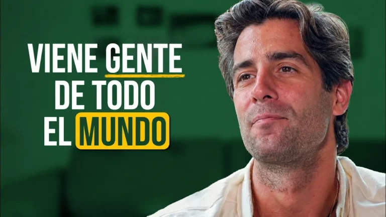 Hombre de pelo canoso y camisa blanca, sonriendo ante un fondo verde con el texto «Viene gente de todo el mundo»