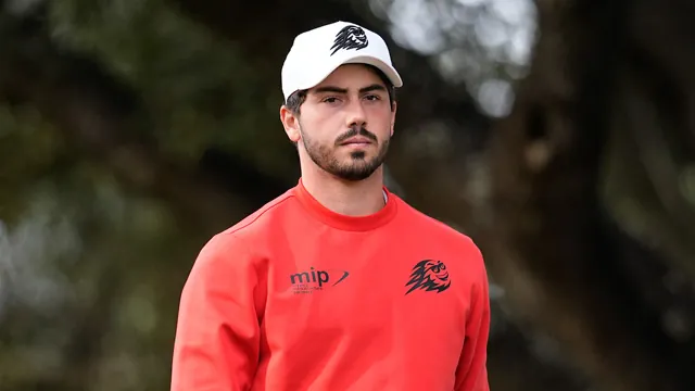 Hombre con gorra blanca y sudadera roja con logotipo, de pie frente a un fondo de árboles desenfocados.