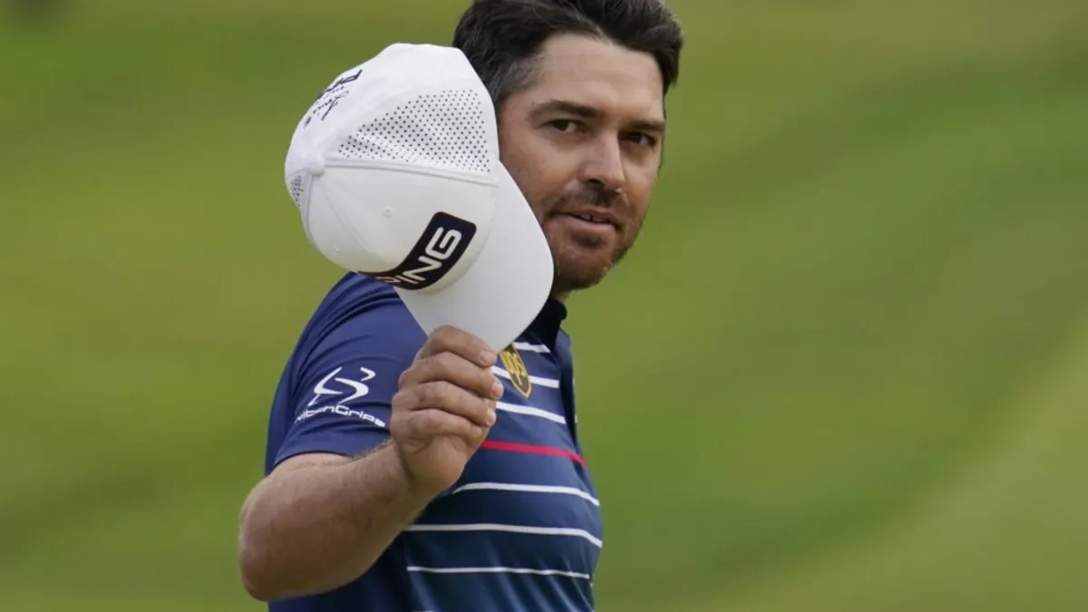 Golfista con polo azul a rayas sostiene su gorra blanca PING y mira a cámara sobre un fondo de césped verde