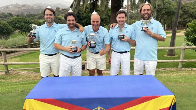 Cinco hombres con polos azul claro sostienen trofeos en un campo de golf, junto a una mesa con mantel azul, amarillo y rojo