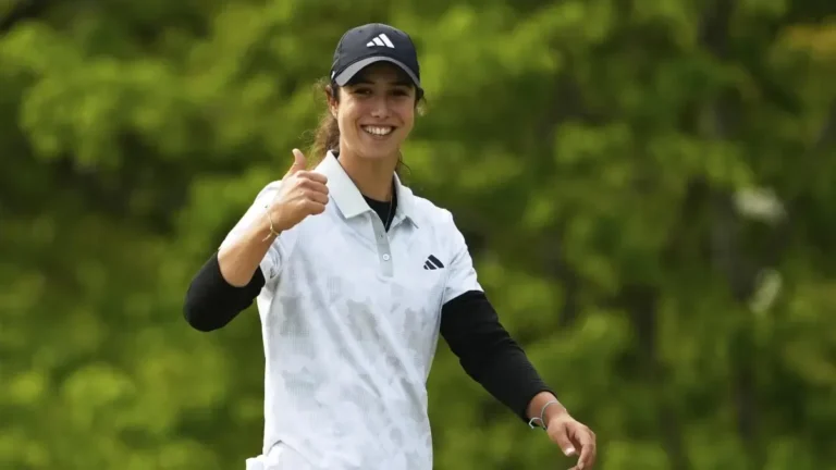 Golfista sonriente con gorra oscura y polo blanco levanta el pulgar ante un fondo de árboles verdes