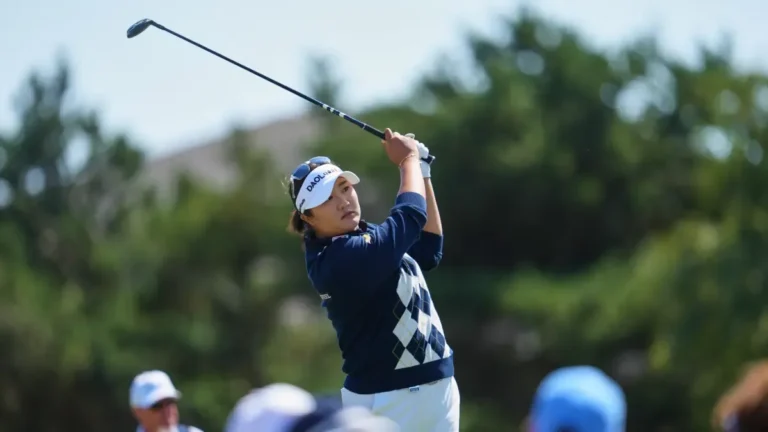 Jugadora de golf con jersey azul marino y visera blanca termina su swing; al fondo, árboles y espectadores.