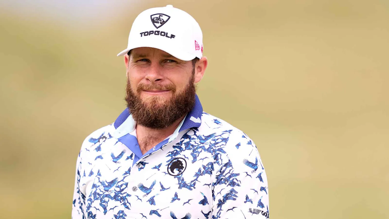 Golfista con gorra blanca de Topgolf y polo con aves azules, sonriendo en un campo de golf desenfocado.