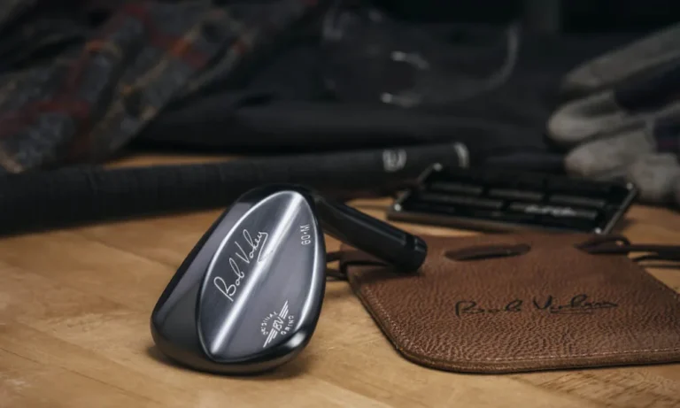 Wedge de golf de acabado negro y cara pulida sobre una mesa de madera, junto a una bolsa de cuero marrón y guantes