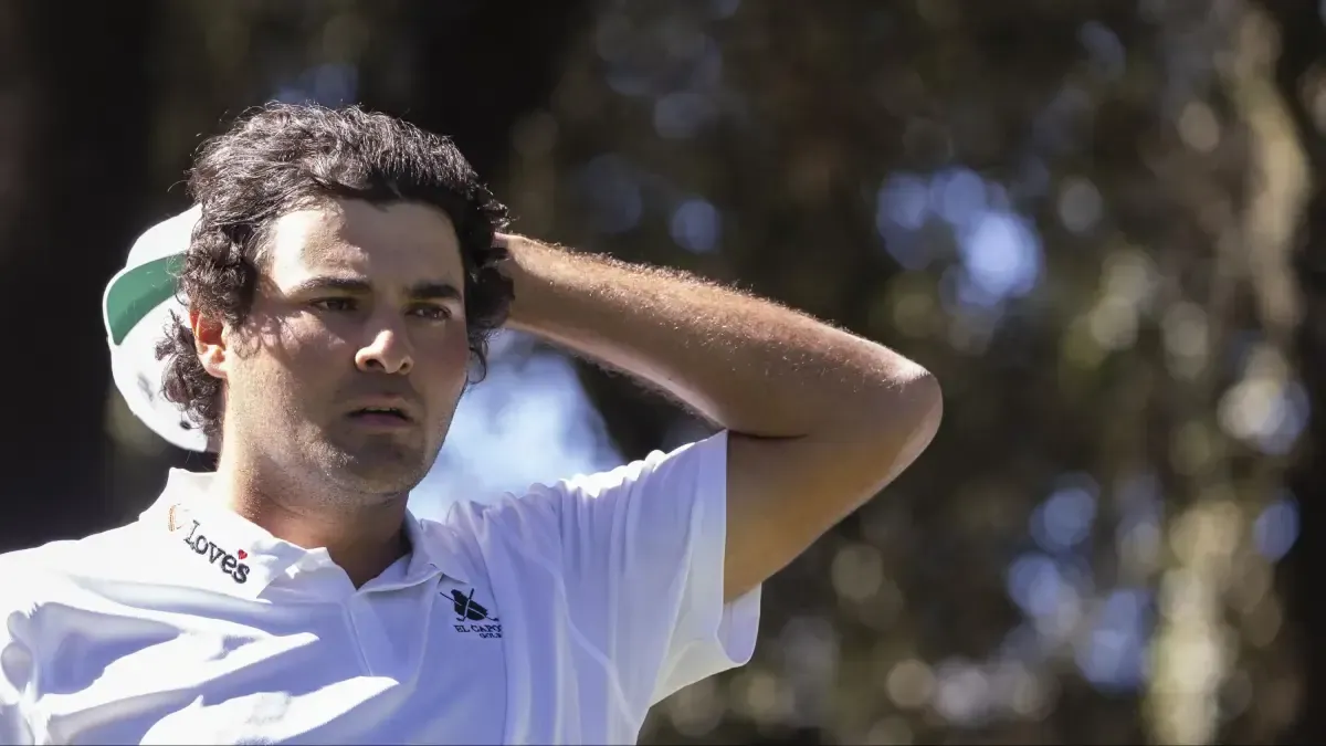 Golfista con polo blanco y gorra verde y blanca, con la mano en la cabeza, mirando a un lado entre árboles desenfocados