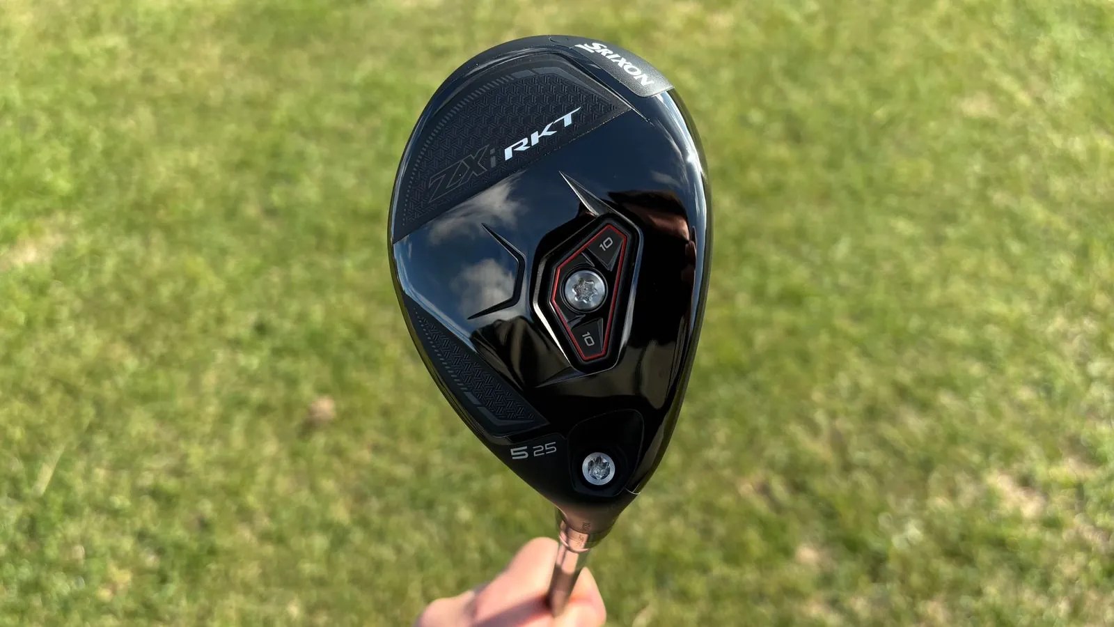 Cabeza negra de un palo híbrido de golf Srixon ZXi RKT, sostenida con la mano sobre un fondo de hierba verde desenfocada.