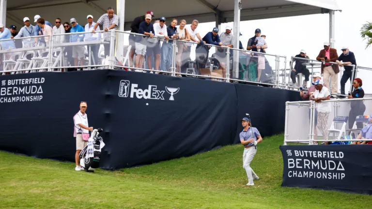 Golfista de polo azul golpea junto a una valla del Butterfield Bermuda Championship, con su caddie y el público de fondo.