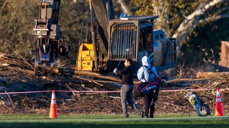 Golfista de jersey negro golpea la bola junto a su caddie; detrás, excavadoras y árboles talados en el campo