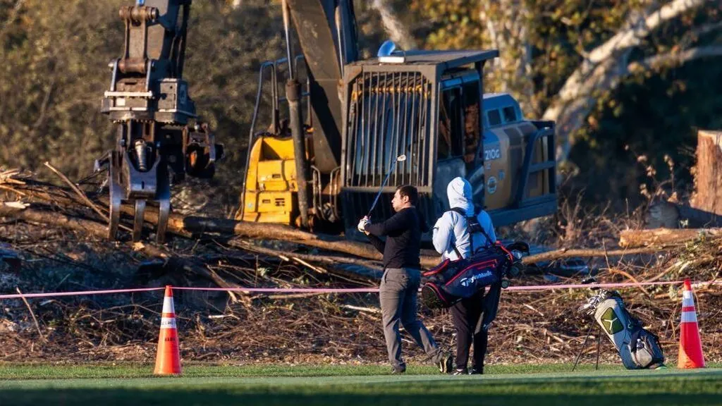 Golfista de jersey negro golpea la bola junto a su caddie; detrás, excavadoras y árboles talados en el campo