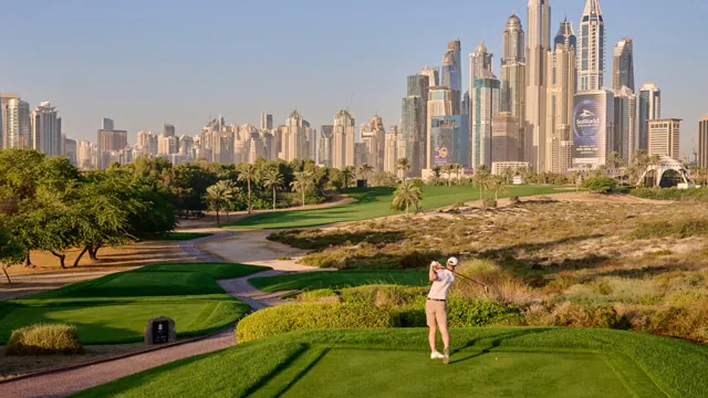 Golfista con gorra blanca y ropa clara golpeando desde un tee con calles verdes, dunas y rascacielos de Dubái al fondo
