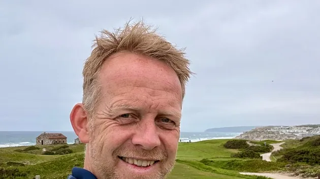 Hombre sonriente en primer plano, en un campo de golf junto al mar, con una casa de piedra y dunas al fondo