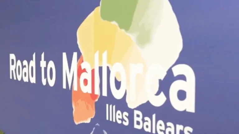 Cartel azul con el texto «Road to Mallorca» e «Illes Balears» y un mapa de Mallorca en colores amarillo, verde y naranja.