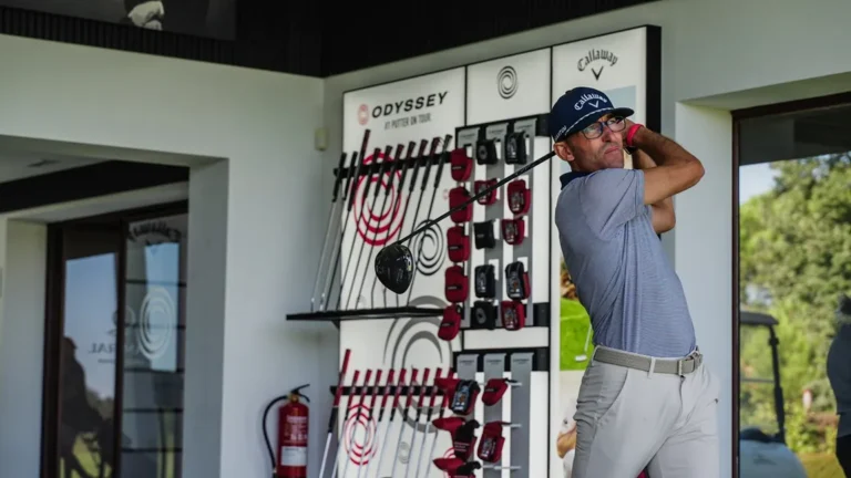 Hombre con gorra azul de Callaway y gafas golpea con el driver en un local de fitting con expositores de palos