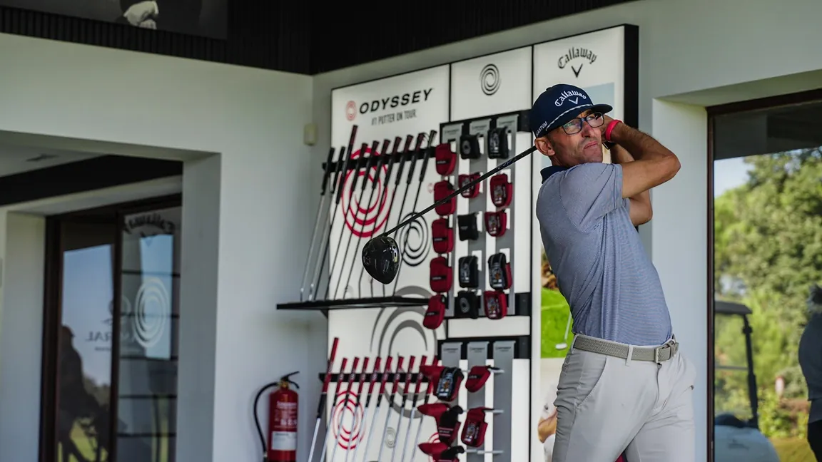 Hombre con gorra azul de Callaway y gafas golpea con el driver en un local de fitting con expositores de palos