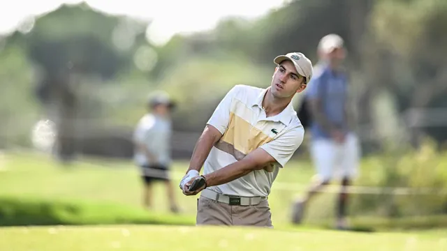 Golfista con gorra y polo blanco a rayas golpeando la bola en un campo de golf soleado, con gente desenfocada al fondo