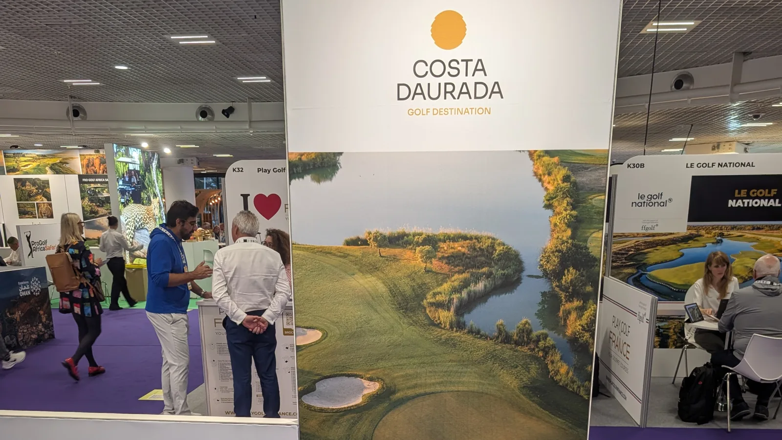 Stand D01 de Costa Daurada Golf Destination en una feria, con un panel vertical con una imagen aérea de un campo de golf junto a un lago, una mesa blanca redonda con un portátil abierto y folletos, y dos sillas blancas