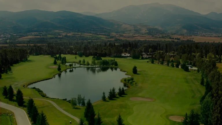 Vista aérea de un campo de golf verde con un lago en el centro, rodeado de árboles y con montañas al fondo